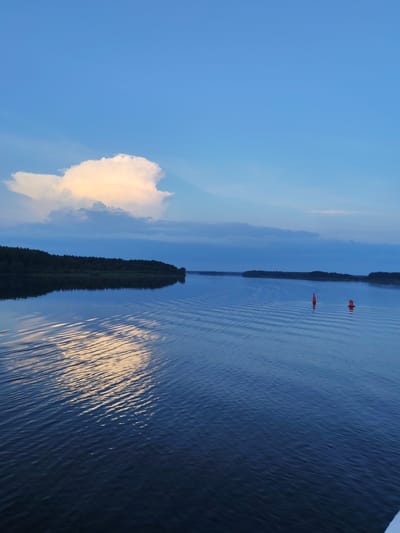 The image depicts a serene body of water, likely a lake or a wide river, under a twilight sky. The water is calm, with gentle ripples reflecting the soft light. Along the left bank, a dense forest of dark green trees stretches horizontally, its reflection appearing sharp and clear on the water's surface. A large, puffy white cloud, tinged with yellow from the setting sun, dominates the upper left portion of the sky, casting a diffused light onto the water below. Further out on the water, two orange buoys are visible, marking a channel or hazard. The overall scene suggests a peaceful, natural environment, possibly on the outskirts of Ul'yanovo, Russia, during the late evening hours. There are no people or any signs of human activity other than the buoys.