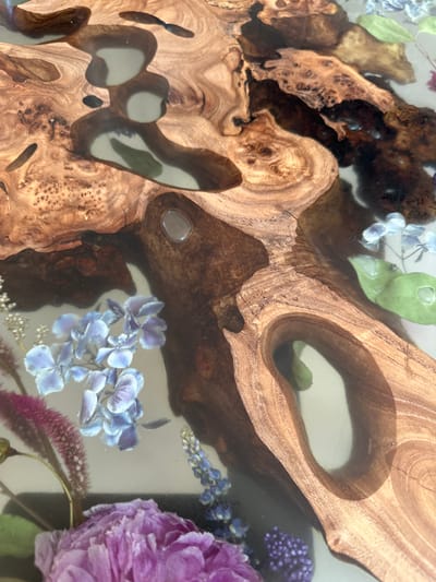 This is a close-up photograph of a resin-encased floral and wood arrangement, likely a tabletop or decorative art piece. The predominant elements are sections of burl wood, with irregular shapes and openings, interspersed with dried flowers in shades of blue, purple, and green. The flowers and wood are embedded in a clear resin. I cannot confirm the location is Малеевка, Russia, without further information.