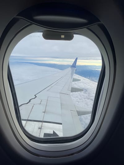 An airplane wing is seen through an airplane window. The wing is white and gray, with visible flaps and rivets. Outside the window, the Earth's surface is visible as a horizon line of blue and tan. Above the Earth, a layer of white clouds fills much of the view. The window has a gray frame and a small gray lever.