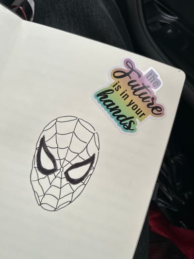 An open white notebook rests on a dark fabric surface, likely a person's leg or lap, in an interior setting located in Trilistnik, Bulgaria. On the notebook's left page, a black ink drawing of Spider-Man's masked face is visible, featuring a web pattern and prominent, solid black eye areas. On the upper right page, an iridescent sticker is affixed, displaying the phrase "the Future is in your hands" in stylized, multi-colored script. The surrounding background consists of dark clothing fabric along the left and top edges, while the right and bottom edges show dark, possibly automotive interior material, with a small patch of red fabric at the lower right corner. No individuals are explicitly visible in the scene.