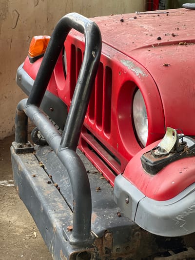 The image is a close-up shot of the front of a weathered red Jeep, likely parked outdoors in Lanžhot, Czechia. The vehicle exhibits signs of disuse, with dust and debris scattered across its hood and grille. A black metal bull bar, rusted in places, protects the front of the Jeep. The right headlight is visible, partially obscured by the bull bar. A broken turn signal housing sits on the right fender. The background suggests an urban or semi-urban environment, possibly a garage or workshop, with a concrete wall and floor visible. No people or actions are depicted in the scene.