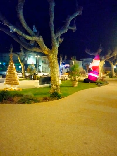 A nighttime scene depicts a town square decorated for Christmas. A white Christmas tree structure displays illuminated place names, with "Feliz Natal" at the base. A large inflatable Santa Claus stands on a patch of grass near a tree. Bare trees frame the scene under a dark blue sky. Buildings and streetlights are visible in the background, adding to the festive atmosphere. The foreground features a paved walkway, bathed in a warm, yellow light.