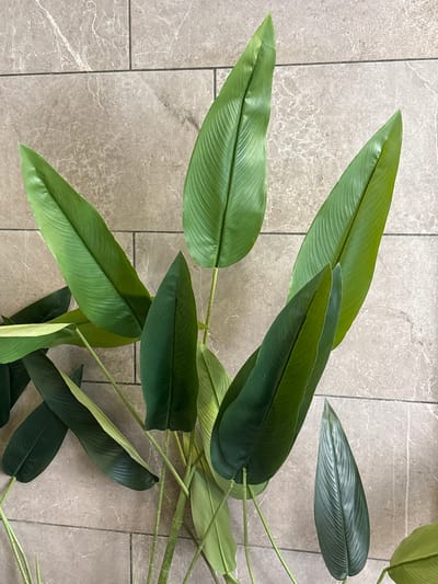 The image displays a collection of artificial tropical plant leaves with long, pointed shapes and prominent parallel veins, arranged against a textured stone tile background. The leaves vary in shades of green, from vibrant lime to deep forest green. The tile background is composed of rectangular tiles with a muted greyish-brown color and a subtle veining pattern. The arrangement of the leaves suggests a decorative display, possibly indoors. The location is identified as Ayn al Basha, Jordan. The scene is static, with no people or actions depicted.