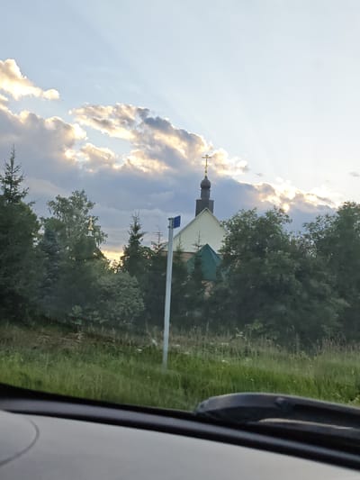 The image shows a church with a gold cross on top, surrounded by trees and a bright sky with clouds. The church is painted white and has a dark, domed roof. There's a green roof section visible below the dome. A light-colored pole with a blue square sign is positioned in front of the church. The foreground shows a grassy area and the edge of a vehicle's dashboard. The scene appears to be set in a rural or suburban area, possibly Ateptsevo, Russia, given the context. The lighting suggests it might be late afternoon or early evening, with sunlight breaking through the clouds.