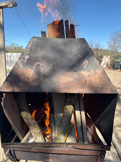 An outdoor scene in Artesa De Segre, Spain, depicts a metal barbecue or fire pit with wood actively burning inside. The primary object is a dark, heat-discolored metal structure, appearing trapezoidal or pyramidal in its upper hooded section, exhibiting areas of black soot and reddish-brown heat tinting and rust. Below the hood, within the firebox, several pieces of wood are burning, characterized by bright orange and yellow flames. The wood pieces include both natural logs with visible bark and lichen, and processed planks. The base of the metal structure shows evidence of rust and wear.

The background features a clear, bright blue sky. The ground surface is an unpaved, light brown dirt or gravel. To the left, a weathered wooden post or support beam is visible, with a thin wire extending diagonally upwards from it. Further left, a white translucent structure, possibly a greenhouse or poly-tunnel, is partially visible. The mid-ground and background contain sparse deciduous trees and brush, along with some indistinct outdoor clutter or storage. No individuals are present in the image.