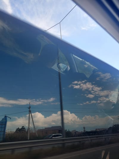 The image captures a view from inside a vehicle, looking out through a side window, with a pronounced reflection on the glass. The picture is rotated approximately 90 degrees clockwise.

**Subjects:** The most prominent subjects are the sky and clouds, which are clearly reflected on the car window. White, fluffy cumulus clouds are scattered across a bright blue sky. Also visible in the reflection are several utility poles with numerous power lines, and a series of buildings or structures, suggesting an urban or suburban landscape along a road. Faintly visible through the reflection and inside the car's window are the silhouette of what appears to be a person's head or a headrest, and part of the car's interior, including a side mirror. The car's interior frame or pillar is visible along the left edge of the frame (top edge when rotated correctly).

**Activity:** The image depicts a scene observed from a car, either stationary or in motion, emphasizing the interplay between the exterior view and its reflection on the window.

**Notable Details:** The time of day is bright daylight, indicated by the clear blue sky and well-defined clouds. The weather appears sunny. The presence of utility poles, power lines, and buildings suggests the vehicle is traveling through a developed area. The strong reflections create a layered visual effect, where the outside world is superimposed with the internal view and environmental reflections. There is no visible text. The overall scene conveys a typical daytime journey.