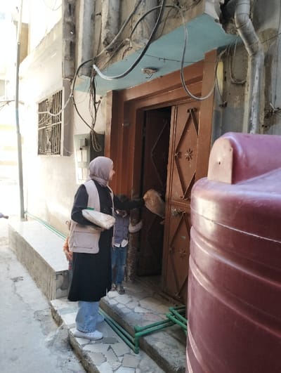 An outdoor scene in قرية الضبية, Syria, depicts a narrow street or alleyway featuring a residential building entrance and several individuals. In the foreground-left, a woman stands facing a partially open brown metal door. She wears a grey hijab, a long black garment, light blue jeans, and white athletic shoes, complemented by a light beige utility vest. She holds a light-colored soft item or bag in her left hand.
To her immediate right, a small child in an orange top is partially visible behind her. Inside the doorway, a second child in blue jeans is partially obscured. The brown double-leaf door features a diamond pattern with a star motif and has a light brown bag-like object hanging from its interior. Above the door, the building facade is light grey with a blue-painted overhang, showing signs of weathering. Numerous tangled black electrical wires run along the upper facade, and a white PVC downpipe is visible on the right. To the left of the door, a barred window is set into the building.
The ground is an uneven mixture of concrete slabs, with a raised concrete curb running along the building's base. Two sets of parallel green metal bars are positioned on the ground directly in front of the doorway. A large, dark red ribbed plastic tank occupies the far right of the foreground, partially obscuring the doorway. Bright sunlight illuminates the scene from the left, casting shadows.