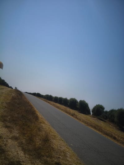 The image depicts a paved road winding uphill through a dry, grassy landscape with a clear blue sky overhead. A line of trees, possibly olive trees, borders the right side of the road, extending into the distance. The dry grass on the left side of the road suggests a warm, arid climate. A light-colored object, possibly part of a hot air balloon or a similar airborne structure, is partially visible in the upper left corner. The scene appears to be a rural or semi-rural setting. The location is identified as Ferreira do Alentejo, Portugal.