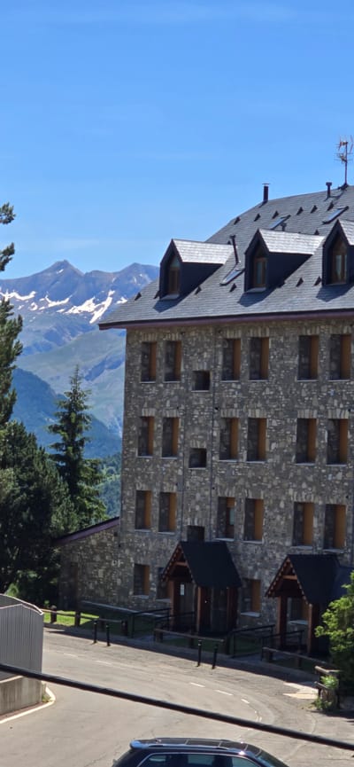 The image depicts a large stone building with a dark tiled roof in Sallent de Gállego, Spain. The building has multiple floors with numerous windows, many of which appear to be dark or boarded up. Several dormer windows with dark triangular roofs are visible on the main building, as well as a smaller stone building attached to its left. The landscape behind the building features lush green trees and majestic, snow-capped mountains under a clear blue sky. A paved road is visible in the foreground, with a dark-colored car parked on it. The scene is a daytime view with bright lighting, suggesting fair weather.