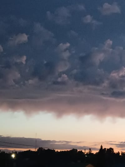 This media file shows a silhouetted landscape against a twilight sky. The upper portion of the image is dominated by a dark blue sky with scattered, wispy clouds. Below this, a band of denser, darker clouds stretches across the horizon, tinged with pink and purple hues. The lower part of the image reveals the silhouettes of trees, buildings, and possibly a transmission tower against a soft gradient of orange, pink, and pale yellow light. A bright, circular light source, possibly a streetlamp or a reflection, is visible in the lower left corner. The overall impression is that of a serene sunset or dusk in a rural or semi-urban setting. The atmosphere appears calm and peaceful. There is no visible text in the image.