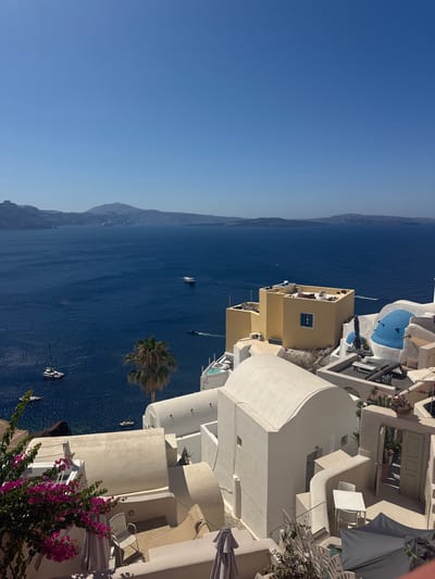 The image captures a picturesque view of Oia, Municipal Unit, Greece, overlooking the Aegean Sea on a clear, sunny day. The foreground is dominated by the iconic white-washed architecture characteristic of Santorini, featuring domed roofs, curved walls, and shaded terraces adorned with potted plants and outdoor furniture. Vivid pink bougainvillea cascades over a railing on the left, adding a splash of color to the monochromatic buildings.
In the middle ground, a bright yellow building stands out, with lounge chairs and air conditioning units visible on its rooftop. Adjacent to it, a section of a building features a striking blue dome and curved white walls. Beyond the structures, the deep blue waters of the Aegean Sea stretch towards the horizon. Several boats, including sailboats and smaller vessels, are scattered across the water. In the distance, a chain of volcanic islands forms a hazy silhouette against the bright blue sky. The scene evokes a sense of tranquility and the idyllic beauty of the Greek islands.