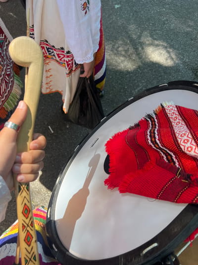 In Bolyarovo, Bulgaria, an outdoor scene is captured from a high-angle perspective, illuminated by direct sunlight creating strong shadows. The foreground features a light-skinned hand, adorned with a plain silver ring on the thumb, gripping a light-colored wooden stick with a curved, hook-like top. The stick's lower visible portion is decorated with red, green, and black geometric patterns. To the right, a large drum with a white head and a black rim is present; the word "MAXTONE" and "THE BEST QUALITY" are visible on its surface. A red, fringed, and intricately woven textile, featuring patterns in red, white, black, and gold, is draped across the drumhead. The shadow of the wooden stick and the hand holding it falls distinctly onto the drum's white surface. In the upper middle ground, the lower torso and arm of an individual wearing a white traditional garment with detailed red, orange, blue, and black embroidered patterns along the hem are visible. The sleeve features delicate white lace trim. This individual's hand holds a black bag. The ground beneath is a textured grey asphalt or concrete surface. Additional colorful, woven textiles, likely part of traditional attire, are partially visible along the left and bottom edges of the frame.
