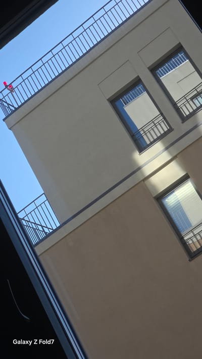 The image shows a low-angle view of a modern building with two visible stories. The building's facade is a neutral beige color, divided horizontally by a dark band. The upper story features two windows with dark frames, each with a small balcony. The windows have internal blinds, and reflections of the sky and other buildings are visible. The lower story has a single visible window, also with a dark frame and internal blinds, and a balcony. A black metal railing runs along the edge of the balconies and the roofline. Above the building, a clear blue sky is visible. On the railing of the upper story, a small red object, possibly a marker or a piece of equipment, is present. The overall scene suggests an urban setting, possibly an apartment complex or office building, with a focus on architectural details and the play of light and shadow. The provided crops highlight specific sections of the building and its surroundings, aiding in detailed observation. The text "Galaxy Z Fold7" appears in the bottom left corner, indicating the device used to capture the image. The location is identified as Krasnoobsk, Russia.