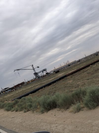 This media file depicts an industrial landscape under a cloudy sky, likely in the Batyr rural district of Kazakhstan. The main subjects are industrial structures and machinery, including a large tower crane and a gantry crane. In the background, a long, low-lying industrial building with a visible power line structure is present. In the foreground, there is a large pipeline running horizontally across the frame, with sparse, dry vegetation and a dirt road. The overall impression is one of an active industrial or construction site, possibly during a construction phase given the presence of the cranes. The time of day appears to be overcast, suggesting it could be morning, afternoon, or evening, with no strong indication of sunlight. The weather is clearly cloudy, contributing to the muted color palette of the scene. There is no visible text in the image. The setting appears to be arid or semi-arid, typical of some regions in Kazakhstan.