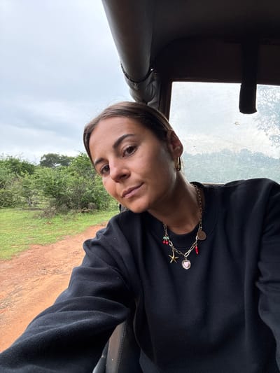 A woman with brown hair pulled back, wearing a black sweater and a gold charm necklace, looks at the camera from inside a vehicle. Visible outside the open window is a dirt path and lush green trees and shrubbery under a cloudy sky. The vehicle's interior is dark, with a partial view of the dashboard and window frame.