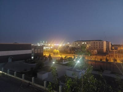 This is an outdoor, eye-level shot taken at dusk or dawn, looking out over a city scene. The sky is a deep, hazy blue, indicating the absence of direct sunlight. In the background, a cityscape is illuminated by artificial lights, with multiple buildings and streetlights visible. One prominent building on the right is a multi-story structure with many windows, some of which are lit from within. Further in the distance, another cluster of buildings appears to be a stadium or sports complex with bright green and white lights.
In the foreground and middle ground, there are more buildings and paved areas. On the left, a large, dark building with a white stripe across its facade dominates the immediate area. Adjacent to it is a paved courtyard with a low fence. Further back, a road runs through the scene, with more streetlights and building lights lining it. There are trees and bushes scattered throughout the landscape, some of which are illuminated by the streetlights, creating lens flares.
There are a few people visible in the lower left portion of the image, walking or sitting near a fence. Their presence is subtle, adding a human element to the urban landscape. The overall atmosphere is quiet and still, characteristic of the transition between day and night. There is no visible text in the image. The location is identified as Mary City, Turkmenistan.