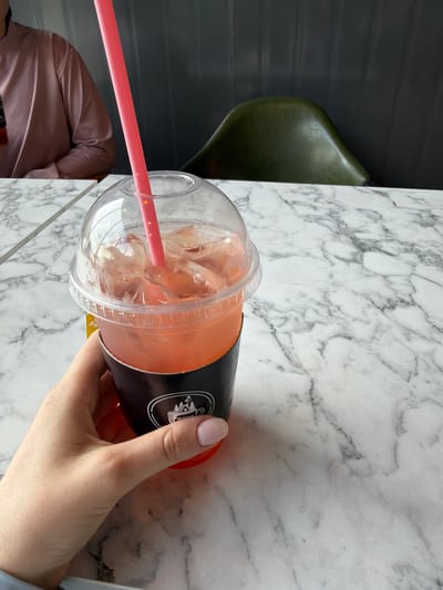 A close-up shot shows a hand holding a clear plastic cup filled with a pink, ice-laden beverage. A pink straw extends from the cup. The cup is encased in a black cardboard sleeve with a white logo featuring trees and a mug. The cup rests on a white marble tabletop. In the blurred background, a person in a pink shirt is visible, and a dark green chair is placed against a dark gray wall. The scene appears to be an indoor establishment, possibly a cafe or restaurant, in Seo-myeon, South Korea.