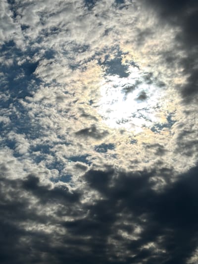A low-angle shot captures a dynamic sky filled with a mix of dark, brooding clouds and bright, luminous clouds illuminated by the sun. The sun is partially obscured by the clouds, creating a radiant halo effect and a sense of depth as light peeks through the gaps. The clouds are textured and varied, with some appearing wispy and others thick and opaque. The overall impression is dramatic and awe-inspiring. There is no indication of the city of Өндіріс, Kazakhstan, or any specific geographical features in this image.