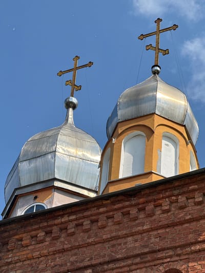The image shows the domes of an Orthodox church in Chudovo, Russia, against a bright blue sky with a few scattered clouds. The foreground is dominated by a brick wall with decorative elements. Two metallic domes, one with a golden cross and another with a similar cross, rise above the wall. The domes are topped with multi-barred crosses, typical of Eastern Orthodox Christianity. The dome on the right is attached to an orange structure with arched windows. The overall scene suggests the exterior of a religious building.