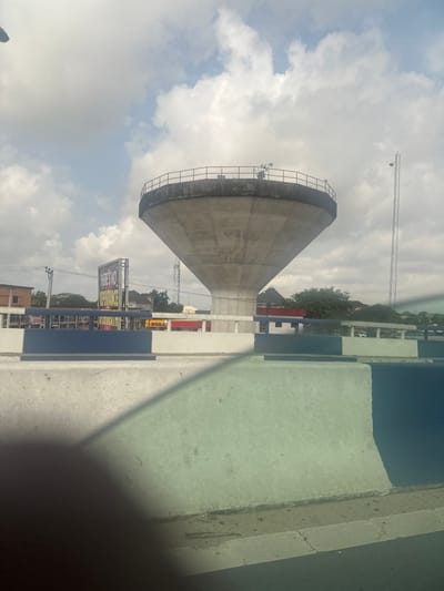 The image depicts a concrete water tower at an unspecified location in Elimgbu, Nigeria, viewed from what appears to be a moving vehicle. The central subject is a large, grey concrete water tower with a wide base that tapers upwards to a cylindrical reservoir section, topped by a circular platform with a dark metal guardrail. A small plant or shrub is visible growing near the guardrail on the platform's right side.

In the immediate foreground, a light grey concrete barrier, possibly part of a road divider or overpass railing, partially obscures the lower portion of the scene. Further back, this barrier transitions to segments painted white and dark blue. A faint green tint and a dark, indistinct shape in the lower left corner suggest the image was captured through a vehicle window, possibly with an obstruction.

To the left of the water tower, a billboard with dark text, partially legible as "THERE'S NO IMPERFECTION," stands in the midground. Beyond the foreground barrier, various low-rise buildings with light and reddish roofs are visible. To the right of the water tower, a tall, slender lattice communications tower is prominent against the sky. The sky is partially cloudy with white and light grey clouds, indicating daytime. No distinct human figures or specific actions are discernible.