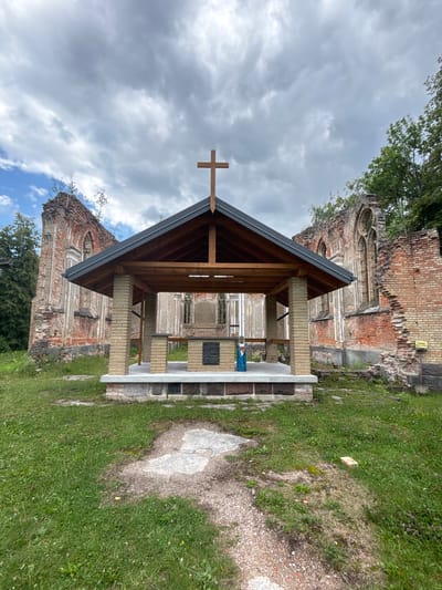 A covered structure with a wooden roof and brick pillars stands in front of the crumbling brick ruins of a gothic church in Bachury, Poland. A wooden cross is mounted on the roof of the covered structure. Inside the structure, a plaque with text is visible, along with a small statue of the Virgin Mary dressed in a blue robe. The ruins are overgrown with vegetation, and the sky above is cloudy. The ground in front of the structure is covered with grass and gravel paths.