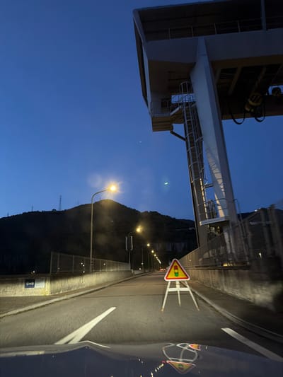 The image depicts a nighttime or early morning scene on the roadway traversing the Barragem de Bagaúste (Bagaúste Dam) near Lamego, Portugal. The view is from the driver's perspective inside a vehicle, with the top of the hood visible in the lower foreground, reflecting the road and a warning sign. The two-lane asphalt road is illuminated by the vehicle's headlights and a series of streetlights extending into the distance on both sides. In the center of the right lane, a portable yellow-and-red triangular warning sign, mounted on a white tripod, displays a black pictogram of a car followed by a second vehicle or trailer.

To the left, a concrete barrier topped with a metal fence runs parallel to the road; an "EDP" (Energias de Portugal) logo is visible on the barrier. Beyond this barrier, a dark, undulating mountain range, dotted with power transmission towers, is silhouetted against a deep blue sky. To the right, a large, light-colored industrial gantry crane structure, integral to the dam's operation, dominates the upper portion of the frame, featuring a control room, support beams, and an external access ladder. The immediate area directly beneath this structure is bordered by a concrete wall and metal fencing alongside the road. The sky is a uniform dark blue, with minimal atmospheric light. No individuals or active interactions are present in the scene.