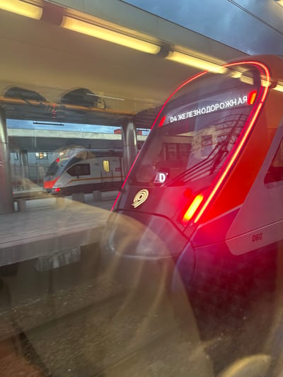 A distorted image shows a reflection of a train at a train station in Aprelevka, Russia. The exterior of a train is orange and gray. The reflection of the train, including its lights, is distorted due to the reflection on glass. There is signage in Russian.