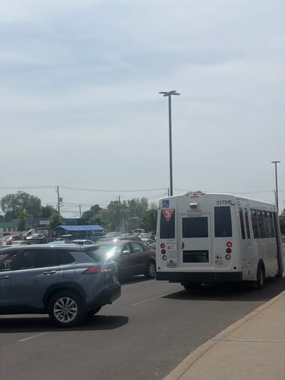 The image captures a bright daylight scene of a white bus and several parked cars, likely in a parking lot or along a street in an urban or suburban area in Longueuil, Canada. The bus is seen from the rear, displaying a strong reflection of light on its metallic surface. Its identifying number "517516" is visible on the upper rear. The emergency door is clearly marked with "PORTE DE SECOURS" in French and "EMERGENCY DOOR" in English. A blue and white sticker with a wheelchair symbol is present, indicating accessibility. Below, there are signs that read "CE VÉHICULE ARRÊTE" (This vehicle stops) and "TRAVERSE À NIVEAU" (at level crossing), accompanied by a red 'X' symbol. The Quebec license plate "AE45148" is affixed to the lower left of the bus.

To the right of the bus, a dark grey SUV is parked, featuring a strong sun flare reflecting off its side. A maroon-colored car is parked behind it. The ground is paved asphalt with a concrete sidewalk or curb running along the right side of the frame. In the background, a line of trees, utility poles, and power lines can be seen. High above, to the left of the bus, an unusual street light or overhead fixture with a cross-like design is silhouetted against the bright, hazy sky. No people are visible in the scene. The strong shadows cast by the vehicles suggest it is mid-day or afternoon.