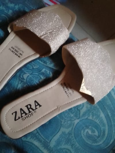 Two glittery, light gold Zara shoes are placed on a blue patterned surface.  The left shoe is partially visible.  "Zara Shoes" is printed in black on the inner sole near the heel. Text indicating "Leather Upper, Man Made Lining, Balance Man Made" is printed closer to the front strap, repeated in a smaller font on the right shoe. The shoes appear to be open-toed, flat sandals with a single wide strap over the foot.