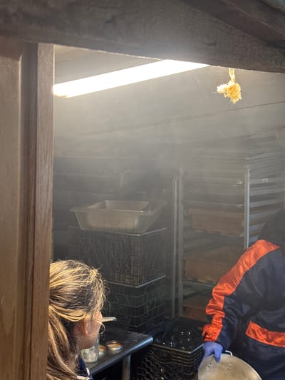 A photograph taken from an elevated vantage point shows a scene inside what appears to be an industrial or commercial kitchen, possibly in Boulder, United States. The overall lighting is a mix of bright artificial light from what appears to be a fluorescent tube and ambient light filtering into the space. The angle suggests the photograph was taken looking down into the area.

A person wearing a dark jacket with orange trim can be seen. They are wearing what appears to be a blue glove on their hand. In their hand, the person is holding a round, flat object. A second person with light brown and blonde hair is seen in the lower right corner of the frame. They appear to be either working in the area or observing the activities.

The room contains a tall metal rack filled with what appear to be flat items. Beneath the rack, there's a metal shelving unit or basket holding various items, including what may be jars or containers. The lower part of the photograph is obstructed by a wooden frame that appears to be the edge of the aperture or structure from which the photograph was taken.

Overall, the scene suggests a busy, functional working environment, likely in a food preparation or storage area.