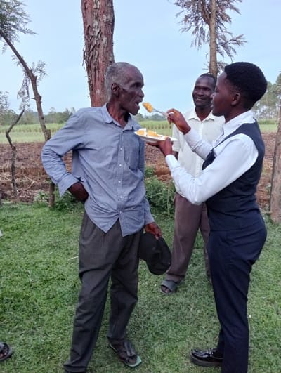 In an outdoor setting, a young woman dressed in a white shirt and dark vest feeds a slice of yellow cake to an elderly man. The man stands with his left hand on his hip and holds a dark-colored hat in his right hand. He wears a blue button-down shirt and dark slacks. Behind them, another man smiles. The scene is set on a grassy lawn with trees and a tilled field in the background.