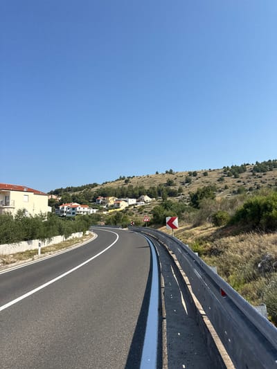 An asphalt road curves through a hilly, Mediterranean landscape under a clear blue sky. On the left, a light-colored building with red-tiled roofs is partially visible behind a line of green bushes. The road is bordered by a concrete retaining wall on the left and a metal guardrail on the right. Several signs are visible along the road, including a yellow warning sign with a red border and a red and white chevron sign indicating a curve. The hillside is covered with dry grass, scattered green bushes, and some trees. In the distance, more houses with red roofs dot the landscape, nestled on the hills. This scene is located in Grad Šibenik, Croatia, a coastal city known for its hilly terrain and Mediterranean climate.