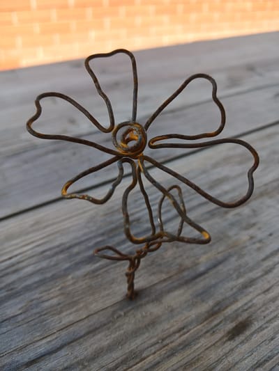 A rusty wire sculpture of a flower with five petals and a twisted stem stands on a weathered wooden surface. The background shows a blurred brick wall with a diagonal shadow. The scene is outdoors, suggesting a yard or patio. The image appears to be a close-up shot, emphasizing the texture and detail of the sculpture. There are no people or animals present. The location is Shud'ya, Russia.