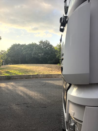 The image shows a close-up, low-angle view of the side of a white truck, parked on an asphalt road. The truck occupies the right side of the frame, with its white body, grille, and headlights visible. In the background, beyond the road and a concrete barrier, is a field of dry, yellowish grass. Behind the field, a dense line of dark green trees forms a natural border. The sky above is partly cloudy, with patches of sunlight breaking through, suggesting it is either early morning or late afternoon. The overall scene appears to be in a rural or semi-rural setting, possibly on the outskirts of Saint-Augustin-des-Bois, France, given the presence of a truck and the natural landscape. There are no people or animals visible in the image. The lighting creates long shadows on the road, indicating the sun is low on the horizon.