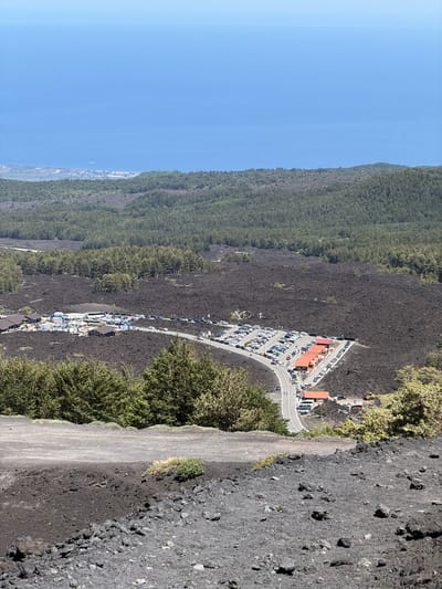 A high-angle, full shot captures the rugged, dark terrain of Mount Etna near Linguaglossa, Italy. The foreground features a steep, dark slope covered in volcanic rock and ash, with sparse patches of green vegetation. In the mid-ground, a paved road winds through a vast expanse of solidified lava flow, marked by dark, uneven surfaces. Along the road, several small buildings with orange-tiled roofs are visible, likely serving as visitor facilities. A large parking lot is filled with numerous cars, and a few tour buses are parked nearby. Beyond the lava field, a dense forest of green trees stretches across the landscape. In the background, the vast expanse of the blue Mediterranean Sea meets the horizon under a clear sky. The scene depicts a popular tourist area on a volcanic mountain, with infrastructure designed to accommodate visitors exploring the unique geological environment.