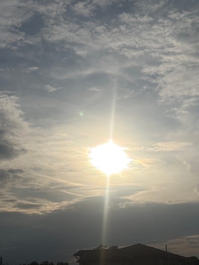 The image captures a vibrant sky scene primarily composed of clouds and a dazzling sun. The sun, located in the upper-middle right section of the frame, is extremely bright, almost blown out, and emits a prominent lens flare that streaks horizontally across the image. A small, faint green orb (a lens artifact) is also visible near the sun.

The sky is filled with a dynamic array of clouds. Near the sun, the clouds are brightly illuminated, appearing white and glowing, while further from the sun, they transition to various shades of grey, indicating depth and shadow. The cloud formations include both wispy, streaky cirrus-like clouds and more textured, lumpy cloud masses, possibly altocumulus or stratocumulus.

On the far right edge of the image, a dark, silhouetted structure, likely a part of a building or a roofline, is visible against the lighter sky. Below this structure, faint outlines of trees or foliage can be discerned, suggesting a terrestrial element in the composition.

The strong, warm glow of the sun, positioned relatively low in the sky, indicates either sunrise or sunset. The weather appears to be partly cloudy with significant atmospheric haze or diffuse light, typical of a bright day with some cloud cover. The scene is set in Kukorevo, Bulgaria. There is no visible text in the image.