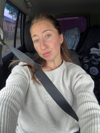 This image depicts a female individual seated inside a vehicle in Karagay, Russia. The subject has fair skin, light-colored eyes (appears blue-green), and light brown hair partially pulled back, with a small cluster of hair visibly caught in the seatbelt. She is wearing a light-colored (off-white or light grey) ribbed knit sweater, which exhibits minor distress or small holes, a small gold-colored hoop earring in her right ear, and a thin silver-colored necklace. Her expression is neutral with her lips slightly parted in a pout, and she is looking directly at the camera. A dark seatbelt crosses her chest and shoulder. Through the left side window of the vehicle, an exterior scene reveals a building with a reddish-brown facade and a prominent blue roof, followed by a smaller, darker building, and sparse vegetation under an overcast or twilight sky. The right rear window of the vehicle shows a purple tint with indistinct shapes in the background. Inside the vehicle, behind the subject, a folded denim jacket and a dark fabric bag with a white and grey circular pattern are visible on the car seats. The car's interior appears dimly lit.