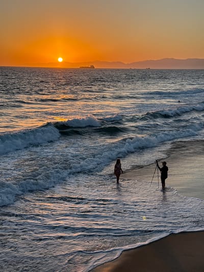 At sunset in Manhattan Beach, California, a man holding a camera on a tripod stands in the shallow ocean surf on the wet sand. A woman in a bikini stands a few feet away in the water, facing the man. The sun is low on the horizon, casting a golden glow across the sky and reflecting on the water. In the distance, a line of hills or mountains is visible, and several ships are sailing on the ocean. Waves are breaking on the shore, creating white foam.