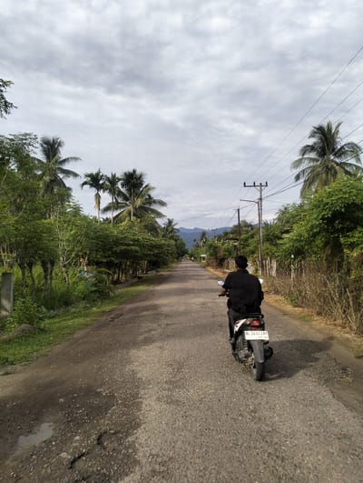 A person rides a black scooter on a paved road in Keudetambue, Indonesia. The rider is facing away from the camera and is wearing a black shirt and pants. The road is lined with lush green vegetation, including palm trees and other tropical plants. In the distance, mountains can be seen under a cloudy sky. Power lines run overhead, supported by wooden poles. The road surface is somewhat worn, with visible cracks and puddles of water.