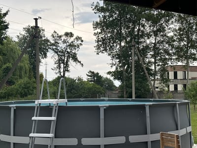 This is a medium-shot, eye-level, outdoor photograph taken on a cloudy day. The foreground features a large, dark gray, above-ground swimming pool with a blue tiled interior. A silver ladder is positioned on the left side of the pool, leading up to the edge. A wooden chair is visible on the right side of the pool. In the background, several tall green trees are present, with a light-colored, multi-story building partially visible behind them to the right. Utility poles with wires are also scattered throughout the background. The overall environment appears to be a residential area with some vegetation and infrastructure.
