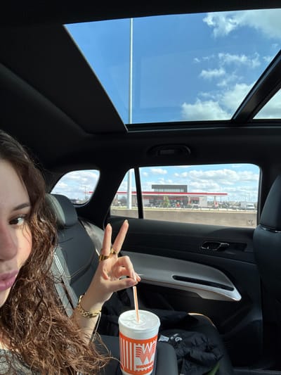 Inside a car with a panoramic sunroof revealing a blue sky with clouds, a person holds up two fingers in a peace sign. They also hold a Whataburger drink with an orange straw. The car interior is black and gray. Outside the window, a large building with red accents is visible. The person has brown curly hair and wears gold jewelry, including rings and a bracelet.