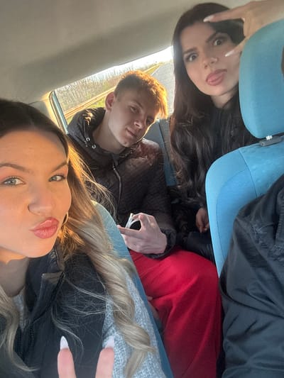 Three individuals are depicted in a selfie-style image taken inside a vehicle. The image appears to have been taken from the perspective of the front passenger seat. The woman in the foreground has long, blonde hair and is wearing a black jacket. The man in the middle has short, light-brown hair and is wearing a black jacket. The woman in the background has long, dark hair and is wearing a blue baseball cap, red pants and black top. The location is presumed to be in Seltsi, Bulgaria. The people are interacting through the photograph. The environment appears to be interior of a vehicle.