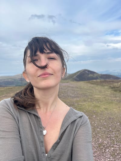 A woman is taking a selfie on a cloudy day with hills in the background. She has dark hair pulled back into a ponytail and is wearing a grey cardigan. A white heart-shaped pendant hangs from a silver necklace around her neck. The landscape appears to be a rural or mountainous area. The specific location in Penicuik, United Kingdom cannot be determined from the image alone.