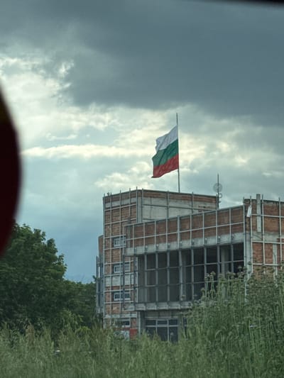 The image shows a building with a steel frame under construction, likely in Пловдив, Bulgaria, as the Bulgarian flag is hoisted on a pole attached to the building's structure. Overgrown vegetation is visible to the right, suggesting the building is on the outskirts of a developed area. The sky is overcast with a mix of dark and light clouds. A red object is partially visible in the foreground, possibly part of a vehicle's interior or exterior.