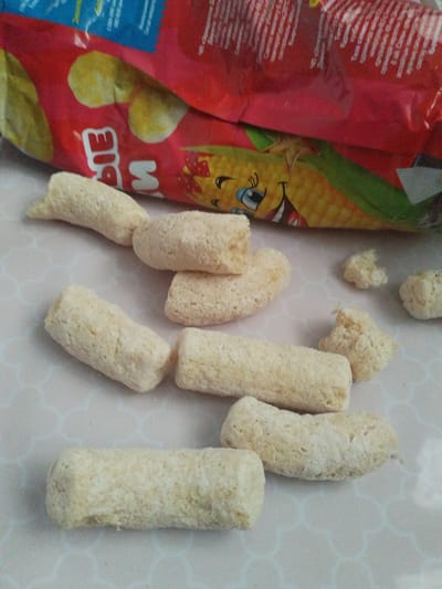 The image shows several light-colored, cylindrical snack pieces scattered on a white surface with a hexagonal pattern. The snack pieces appear to be puffed corn snacks, some intact and others broken. In the background, a portion of a red and yellow snack bag is visible, featuring graphics of corn and a cartoon character. The scene does not provide any specific indicators related to the city of Октябрь, Kyrgyzstan, or any geographical context.