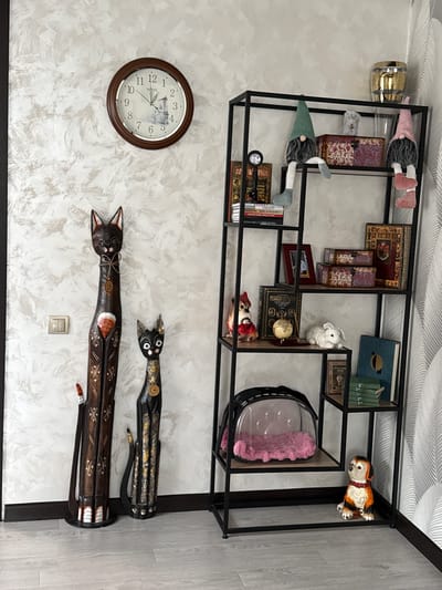 An indoor scene showcases a decorative wall display in Krapivka, Russia. A large black metal open-frame shelving unit is mounted on a light-colored, textured wall. The shelves are adorned with various decorative items, including several gnomes with soft bodies and pointed hats (one in pink, one in green), a small white knitted rabbit, and ceramic figures of a dog and a small cat/fox in a red hat. Numerous books are arranged on the shelves, some with ornate covers in dark red and gold, others in blue and green. Two decorative chest-like boxes with patterned fabric, a small globe, a framed picture, and a clear container holding a pink fluffy item also populate the shelves. A gold-colored lamp fixture is visible near the top left shelf.
Below the shelving unit, a round wall clock with a dark brown frame and a white face showing a landscape picture marks the time at approximately 11:36. The brand "SINIX" is visible on the clock face. To the right of the clock, two long, stylized carved wooden cat figures, dark brown with gold, white, and red embellishments, are mounted on the wall. A light switch is positioned below the wooden cats. No activity or event is taking place in the static display.
Visible text includes: "SINIX" on the clock, and "Гурва" on the spine of one of the books.