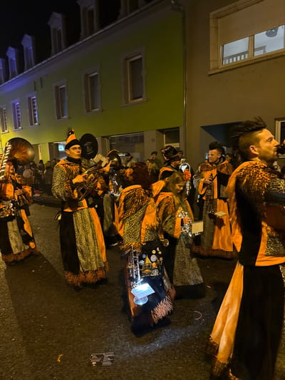 The image depicts a nighttime street scene in Wasserbillig, Luxembourg, featuring a group of individuals, likely a brass band or carnival procession, dressed in elaborate costumes. The attire consists primarily of orange and black fabrics with leopard print patterns and faux fur trim.

In the foreground, a prominent male figure on the left wears a pointed orange and black hat with a feather plume, spectacles, and holds a euphonium. To his left, a partial view of another individual shows them holding a large tuba. Central to the frame, a person with their back mostly to the camera wears a similar costume, including a dark apron-like garment adorned with pins and badges, carrying what appears to be a small, illuminated instrument case.

Further to the right, a man with dark, spiked hair is seen in profile, raising a dark bottle to his lips. Other similarly costumed individuals are visible throughout the mid-ground, some holding brass instruments, including trumpets or cornets. In the background, additional people and vague shapes of percussion instruments can be discerned.

The setting features buildings lining the street. The building on the left has a light green facade with multiple windows featuring white frames and several dormer windows on the roofline. A storefront or commercial entrance is visible at ground level. The building immediately to its right has a grey facade with at least one window featuring a closed roller shutter. The ground is dark asphalt, and some scattered debris, including a small piece of paper or card and a pink rubber band, is visible underfoot. The scene is illuminated by artificial street lighting, casting shadows and highlighting the subjects against the dark environment.