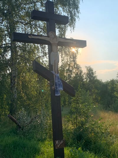 A dark wooden crucifix, depicting Jesus Christ, stands in a grassy field with trees in the background. The sun is setting behind the crucifix, casting a warm glow and creating lens flare. A white cloth with blue embroidery is tied to the crucifix. At the base of the crucifix, a small, worn symbol is visible. In the foreground, green grass and some dry vegetation are present. In the mid-ground to the left, a metal pipe is leaning against some bushes. The sky is clear with a few wispy clouds, and the overall impression is one of a rural, serene setting. The image was taken in Mala Rublivka, Ukraine.