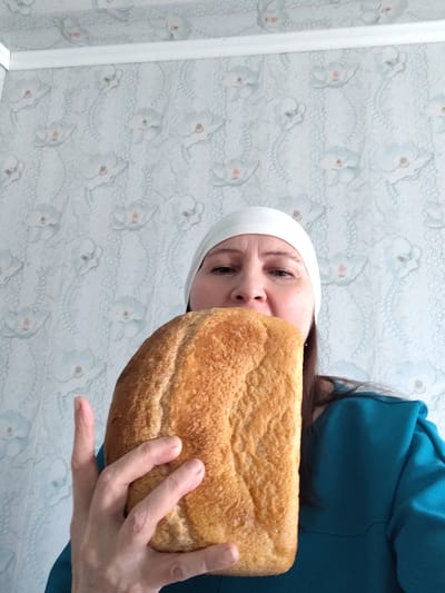 A medium shot captures a woman indoors, holding a large, golden-brown loaf of bread in front of her face. Her eyes and forehead are visible above the bread, and she looks directly at the camera with a neutral expression. She wears a plain white head covering, possibly a cap or bandana, and a teal-colored long-sleeved top or dress. Her right hand is visible holding the bread from below.
The background consists of a light-colored wallpaper featuring a repeating pattern of white orchids and delicate, light blue lace-like motifs. A white decorative trim or molding runs horizontally across the top of the wall, suggesting the transition to a ceiling. The setting is clearly a domestic interior. The lighting appears to be soft, ambient light, indicative of daytime indoors. There are no strong shadows or other weather indicators.
The activity shows the woman holding the bread close to her face, partially obscuring her mouth and nose. This could imply she is presenting the large loaf, possibly about to consume it, or engaging in a playful interaction with the food. No text is visible in the image. The overall scene suggests a casual moment within a home in Сусады-Эбалак, Russia.