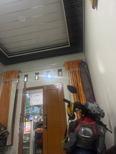 The image depicts an indoor scene, likely a room within a residential property in Serdang Kulon, Indonesia, as suggested by external context. The primary focus is a red and black motorcycle parked in the foreground. The motorcycle has a black handlebar with a yellow item and a grey cloth draped over it. Behind the motorcycle, a wooden door stands slightly ajar, revealing a glimpse of an outdoor area with greenery. To the left of the door, orange curtains with white lace trim hang over a window. Further left, a black box with "BUFF" branding is visible.
The room's ceiling is a modern design, with white horizontal panels framed by black sections featuring a wavy pattern. Several rectangular windows with wooden frames are set into the wall above the door and curtains, with light emanating from them. A large, rolled-up rug with a floral pattern is propped against the wall to the right of the curtains. The walls are painted white. The overall impression is that of a functional interior space.